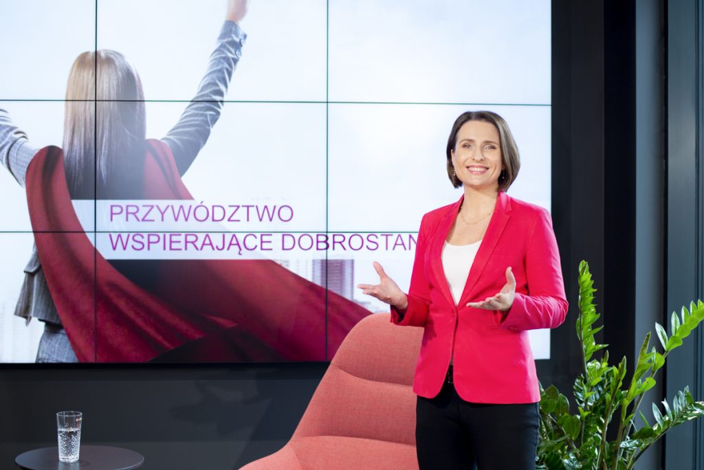 Kobieta w czerwonej marynarce prowadzi prezentację. Za nią na ekranie widać napis "PRZYWÓDZTWO WSPIERAJĄCE DOBROSTAN".